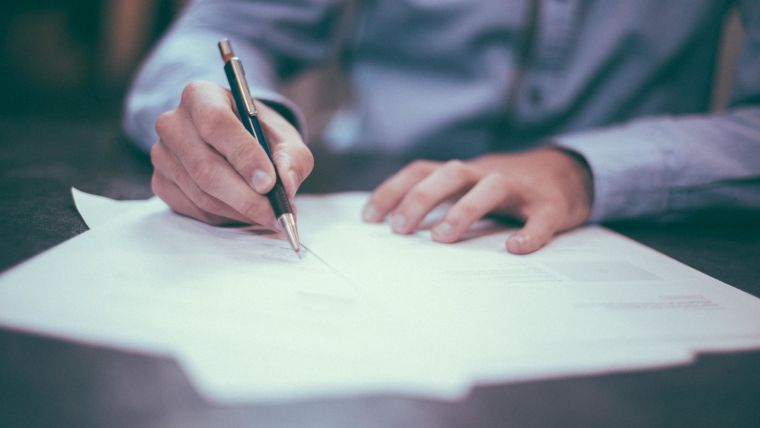 Stock image of a person in a blue shirt writing on paper with a pen.